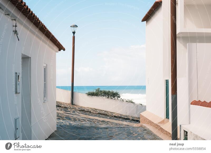 Blick auf den Meereshorizont zwischen zwei Häusern – Zentralperspektive Küste Dorf weiß blau Sommer freundlich Idylle Ruhe entspannung Weg Steigung Straßenlampe