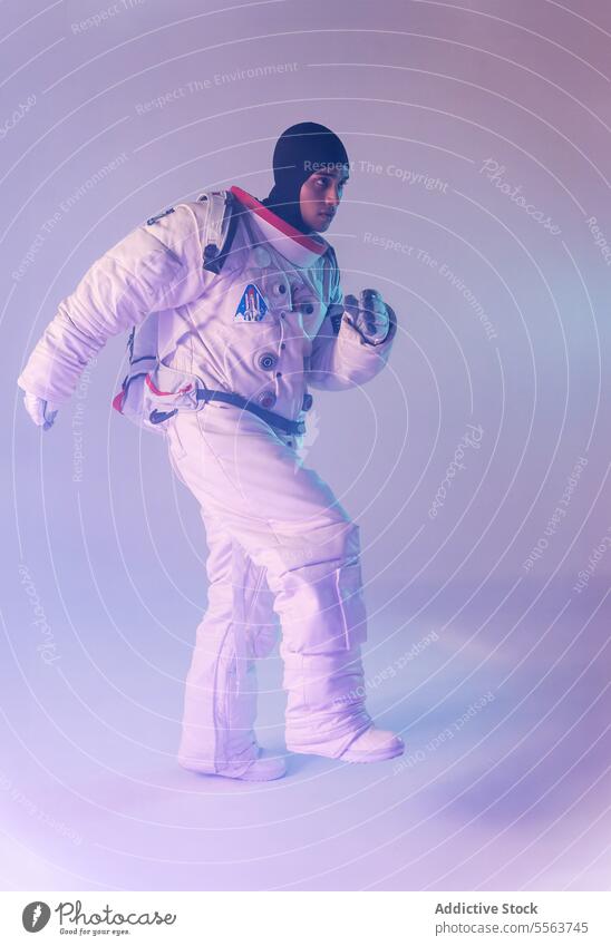 Männlicher ethnischer Astronaut in Raumanzug und Rucksack mit dunkler Kapuze marschiert vor lila Hintergrund Kosmonaut erkunden Schmuckkörbchen Astronomie