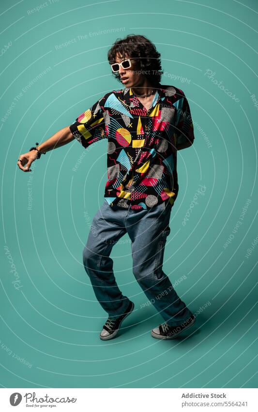 Stilvoller junger ethnischer Mann tanzt mit bewegten Beinen und Armen in der Nähe einer türkisfarbenen Wand flippig Tänzer ausführen aufgeregt sich[Akk] bewegen