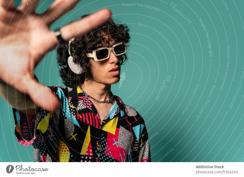 Trendiger ethnischer junger Mann mit Kopfhörern und Sonnenbrille vor türkisfarbenem Hintergrund zeigen farbenfroh gestikulieren Hemd flippig Mode zuhören
