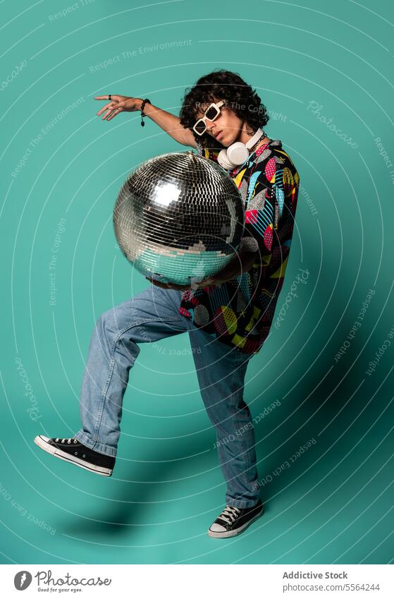 Synkopierter junger ethnischer Mann tanzt mit Discokugel mit erhobener Hand und Bein Sonnenbrille zeigen farbenfroh gestikulieren Hemd flippig Mode