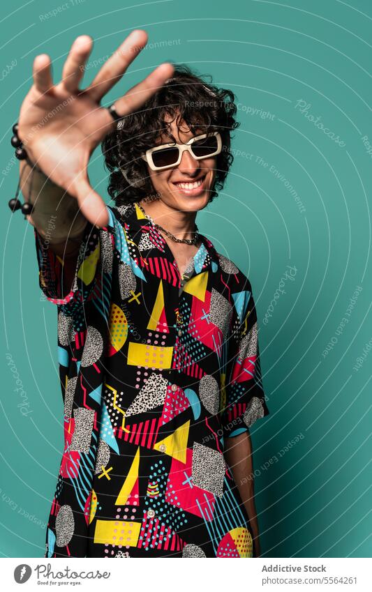 Trendiger ethnischer junger Mann mit Kopfhörern und Sonnenbrille vor türkisfarbenem Hintergrund zeigen farbenfroh Lächeln gestikulieren Hemd flippig Mode