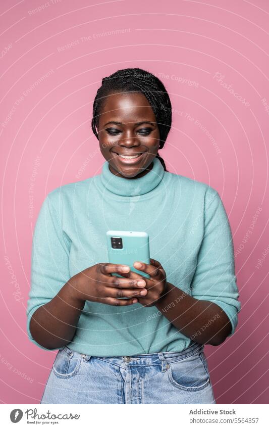 Afrikanische Frau lächelt auf ihr Smartphone in einer rosa Umgebung blaugrün Rollkragenpulli türkis Hintergrund Freude Stil Mode Porträt Technik & Technologie
