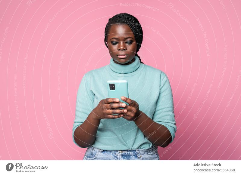 Fokussierte afrikanische Frau mit Telefon vor rosa Hintergrund türkis Rollkragenpulli Smartphone hell-blau fokussiert jung Zopf Mode Stil modern