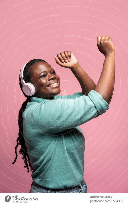 Fröhliche Afrikanerin mit Kopfhörern Afrikanisch Frau rosa Hintergrund Freude Musik zuhören anheben Hand Lächeln Jugend Emotion Fröhlichkeit feiern Tanzen