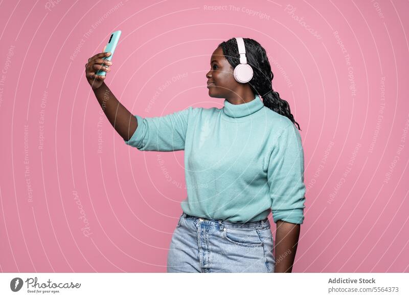 Afrikanische Frau genießt Musik vor einem rosa Hintergrund Kopfhörer Smartphone blaugrün Pullover zuhören Audio Entertainment Profil Freizeit Zopf Gerät Hand