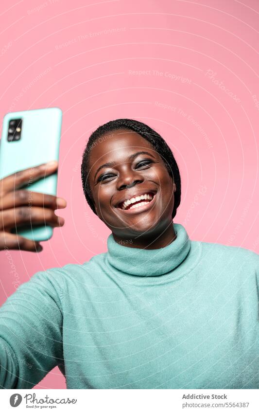 Fröhliche afrikanische Frau nimmt ein Selfie vor rosa Hintergrund Nahaufnahme Gesicht Afrikanisch Lächeln Smartphone blaugrün Emotion Freude Auge Make-up