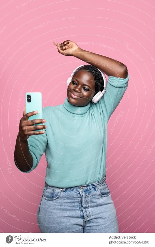 Afrikanische Frau macht Selfie mit Smartphone vor rosa Hintergrund Kopfhörer Tanzen heiter Freude Mobile Telefon Spaß Musik Rhythmus feiern Porträt pulsierend