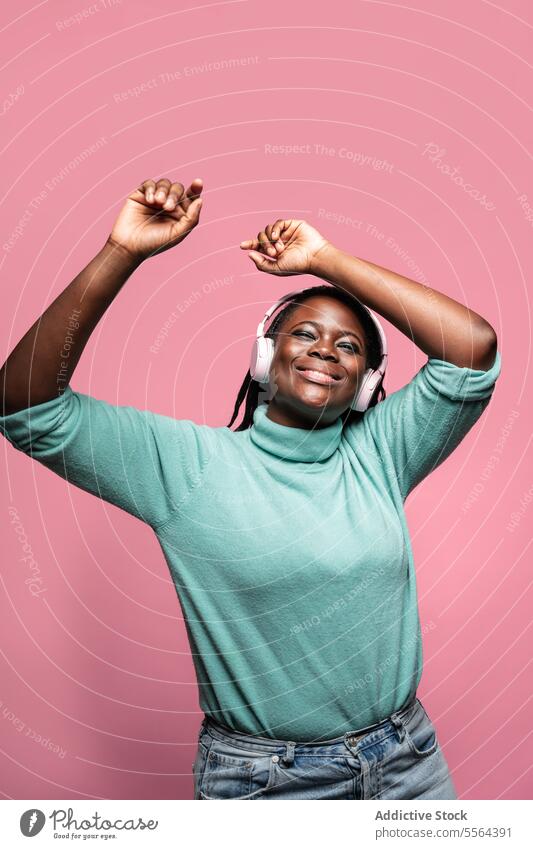 Fröhliche Afrikanerin mit Kopfhörern Afrikanisch Frau rosa Hintergrund Freude Waffen angehoben Tanzen Musik Fröhlichkeit grün Top Entertainment Feier Audio