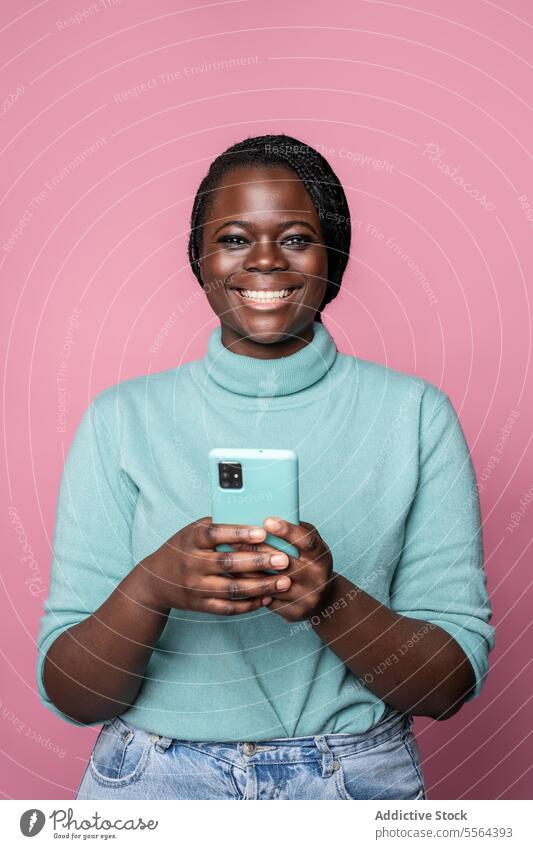 Afrikanische fröhliche Frau mit Smartphone und blauem Pullover vor einem rosa Hintergrund heiter türkis Rollkragenpulli hell-blau Lächeln breit jung Zähne Zopf