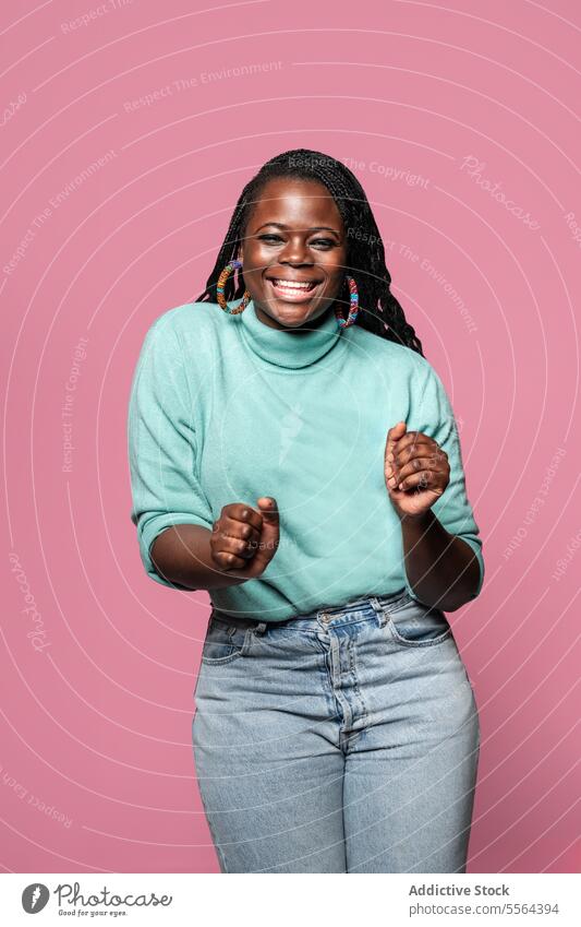 Fröhliche Afrikanerin im türkisfarbenen Pullover Afrikanisch Tanzen Frau Jeanshose Bewegung rosa Hintergrund Lächeln Freude Aufregung Faust pressen Jeansstoff