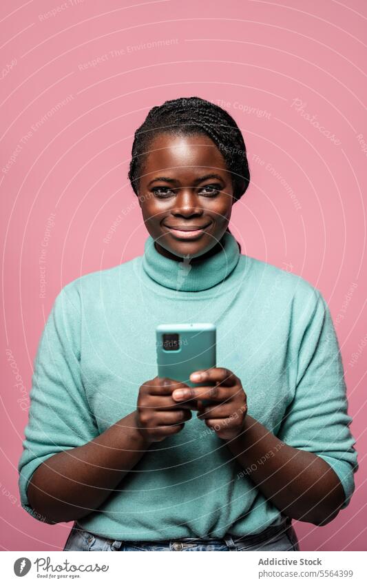 Porträt einer lächelnden Frau mit Smartphone Afrikanisch Lächeln blaugrün Pullover rosa Hintergrund heiter Halt Freude Mitteilung Mobile Technik & Technologie