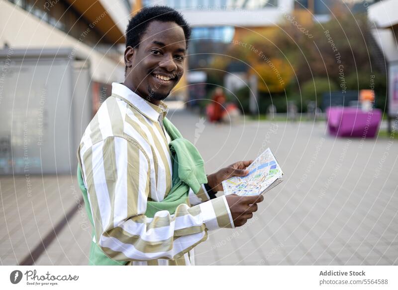 Fröhlicher schwarzer junger Mann steht mit einer Landkarte auf einer gepflasterten Straße bei Tag Gebäude Glück Lächeln Großstadt heiter männlich stehen