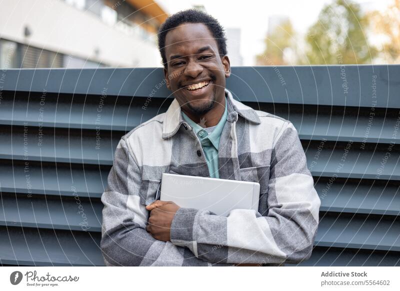 Fröhlicher junger schwarzer Mann steht mit Laptop im Park bei Tag Lächeln Porträt Glück heiter Gebäude positiv Großstadt die Arme verschränkt Sommer männlich