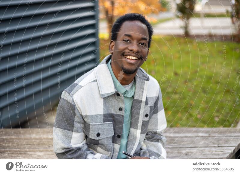 Fröhlicher junger schwarzer Mann auf einer Bank sitzend mit Stift und Kugelschreiber in der Tageszeit Schreibstift Lächeln Porträt Glück positiv Gebäude
