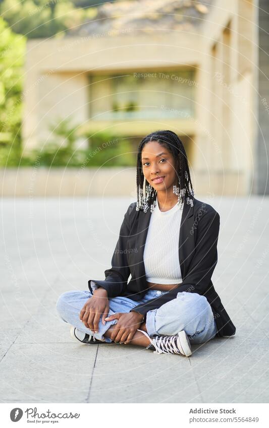 Lächelnde afroamerikanische Frau sitzt auf dem Boden im Hof eines Gebäudes Vorschein selbstbewusst lässig Porträt Glück Stil Afro-Look positiv Geflecht sitzen
