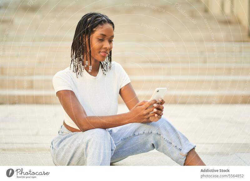 Junge ethnische Frau, die auf einer Treppe sitzend ihr Mobiltelefon benutzt Smartphone benutzend Browsen Treppenhaus Glück Mobile Telefon Surfen Lächeln Schritt
