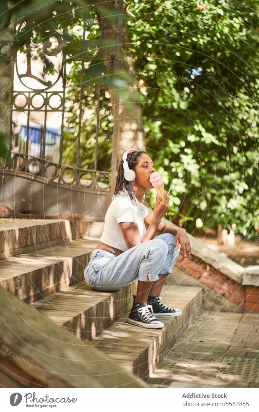 Schwarze Studentin mit Kopfhörern und einer Eistüte auf einer Treppe sitzend Frau Schüler Löffel Speiseeis Campus Zapfen Treppenhaus Sommer jung ethnisch