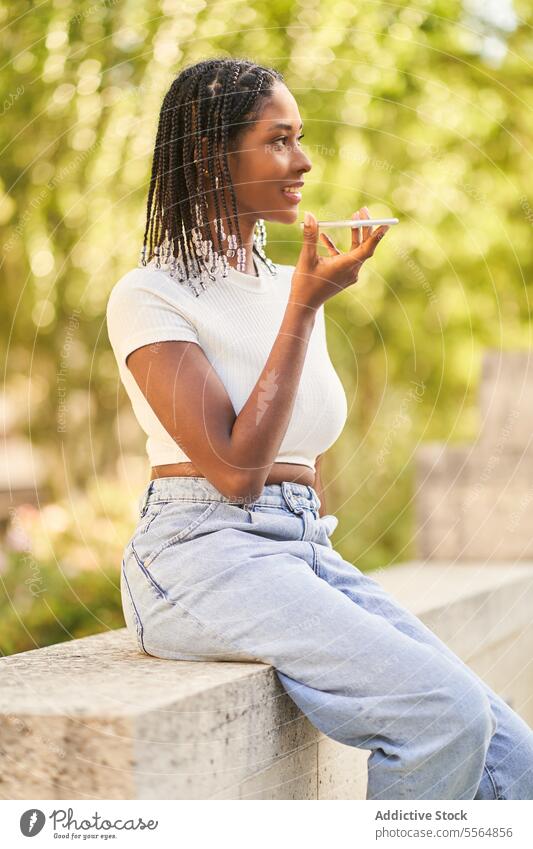 Afroamerikanische Frau, die auf einer Steinbank sitzt und mit ihrem Smartphone eine Sprachnachricht aufnimmt Nachricht Audio Stimme benutzend Borte Park