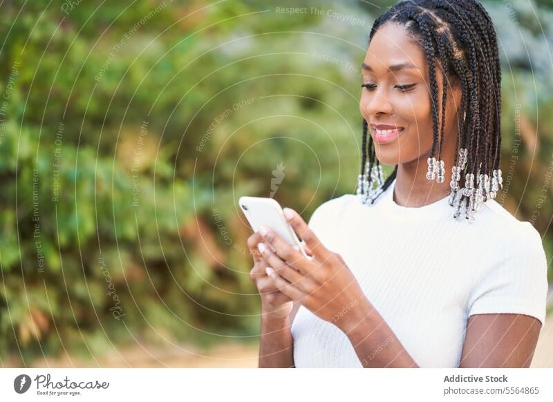 Fröhliche ethnische Frau, die auf der Straße in der Nähe von grünen Büschen und Bäumen auf ihrem Smartphone surft benutzend Glück Telefon Browsen Lächeln Mobile