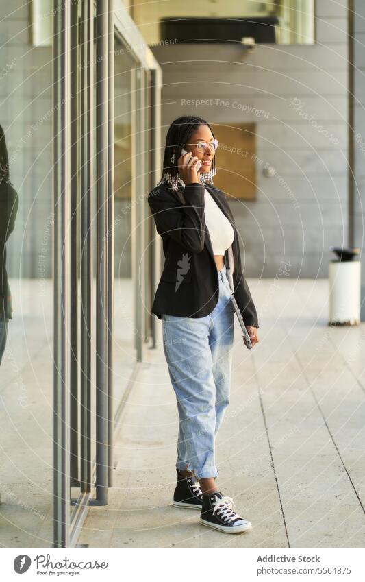 Lächelnde Geschäftsfrau, die spazieren geht und mit dem Mobiltelefon spricht, während sie einen Laptop hält Frau Smartphone Spaziergang Eingang sprechen
