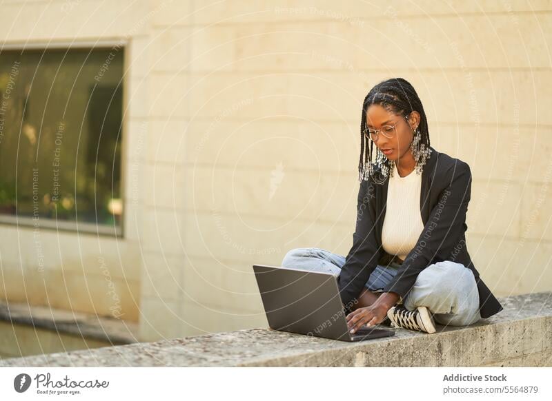 Konzentrierte ethnische Geschäftsfrau, die an einem Laptop arbeitet, während sie an einem Steinrand in der Nähe von Pflanzen sitzt Frau Arbeit freiberuflich