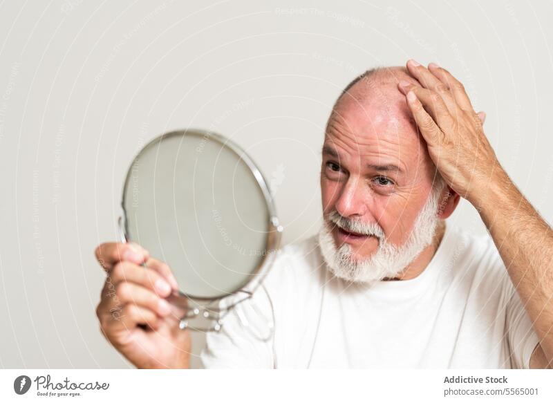Älteres männliches Modell mit Glatze betrachtet den Spiegel im Studio Mann Hautfalten haarlos kahl Vorschein Teint Pflege Selbstversorgung Routine täglich