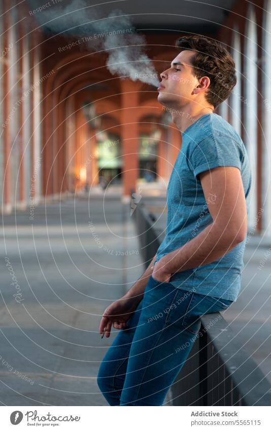 Junger Mann beim Ausatmen von Rauch in der Nähe von Torbögen in einer Stadt. Kaukasier ausatmend Gen-Z Reling Bögen jung Lehnen Jeanshose Hemd im Freien Gebäude