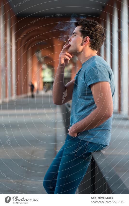 Junger Mann raucht in der Stadt und sitzt in einem städtischen Geländer. jung Rauchen Großstadt Reling ausatmen Gen-Z Stehen blau T-Shirt Jeanshose Sitzen