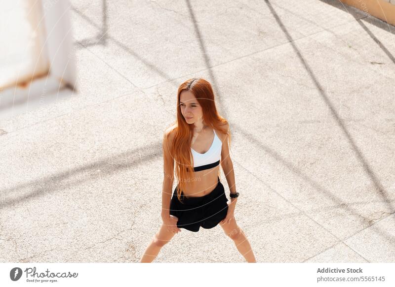 Junge rothaarige Frau streckt sich auf der sonnenbeschienenen Straße Rotschopf strecken im Freien sonnig Tag Sport-BH schwarze Shorts Straßenbelag Fitness Übung