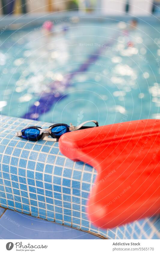 Schwimmbrille und Kickboard am Beckenrand mit verschwommenem Kind Pool im Innenbereich Schutzbrille rot blau Fliesen u. Kacheln schwimmen Wasser Gerät Sport
