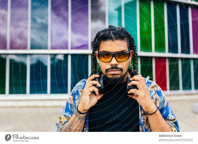 Lateinamerikanischer Mann, der Kopfhörer vor einem bunten Gebäudefenster einstellt. Zopf Beteiligung Sitzen Sonnenbrille Musik Hintergrund Vielfalt im Freien