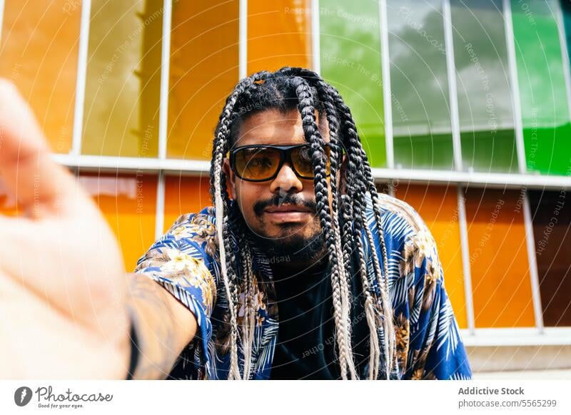 Ein lateinamerikanischer Mann macht ein Selfie vor lebendigen Fenstern. Latein Geflecht Sonnenbrille Smartphone Ästhetik farbenfroh alternativ Gesicht Porträt