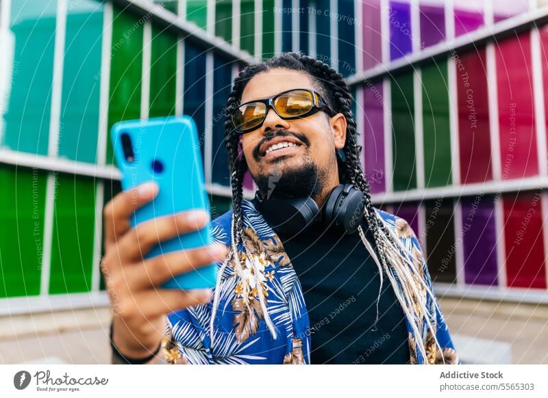 Lateinamerikanischer Mann mit Kopfhörern im Nacken, der auf ein blaues Smartphone in einem bunten städtischen Raum schaut. Zopf Lächeln sprechend farbenfroh