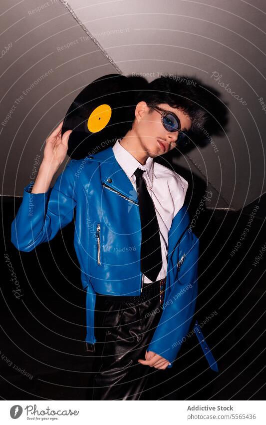Mann im Retro-Look mit Schallplatte im Dunkeln stylisch Brille faulenzen Vinyl Aufzeichnen blau Jacke Leder Hose dunkel retro Musik Mode Stil 80s Ambiente
