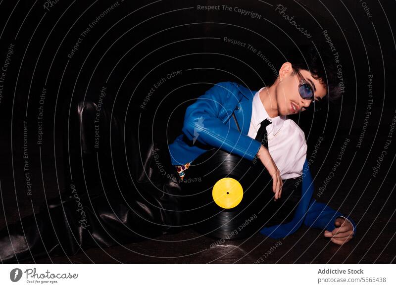 Mann im Retrostil mit Schallplatte im Dunkeln lümmelnd stylisch Brille faulenzen Vinyl Aufzeichnen blau Jacke Leder Hose dunkel retro Musik Mode Stil 80s