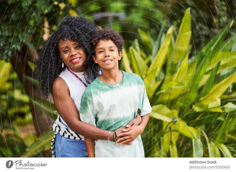 Freudige Umarmung von Mutter und Sohn in tropischer Umgebung Frau Junge Umarmen Pflanzen Freude Lächeln krause Haare Liebe Familie Zuneigung grün Hemd binden