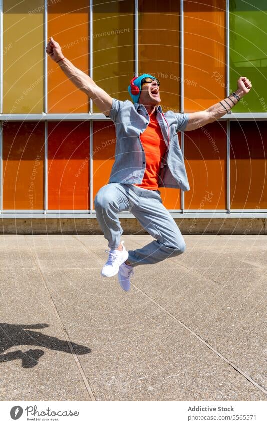 Energiegeladener Mann feiert mit Sprung vor bunter Kulisse springen Feier Kopfhörer Musik Freude lässig pulsierend Farbe Gebäude Hintergrund Architektur