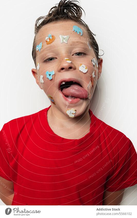 Junge mit bunten Aufklebern im Gesicht und nassen Haaren Zunge farbenfroh Tier nasses Haar spielerisch Kind jung Ausdruck rotes Hemd weißer Hintergrund Spaß
