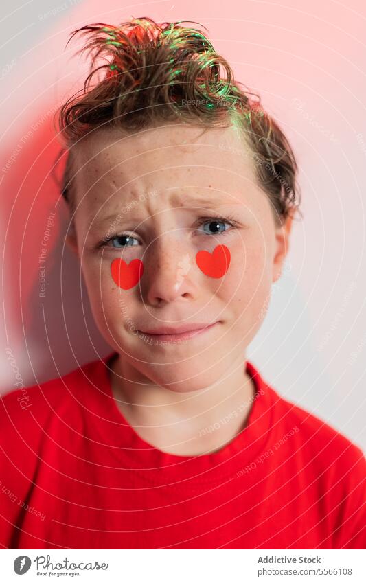Ruhiger Junge mit Herzaufklebern auf den Wangen und abstrakten roten Mustern Aufkleber krause Haare Windstille Starrer Blick rotes Hemd weißer Hintergrund