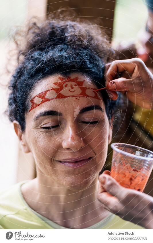 Lächelnde Frau mit geschlossenen Augen malt auf Gesicht Farbe Augen geschlossen Tradition Glück Kunst Lebensmitte krause Haare positiv Stirn froh Optimist