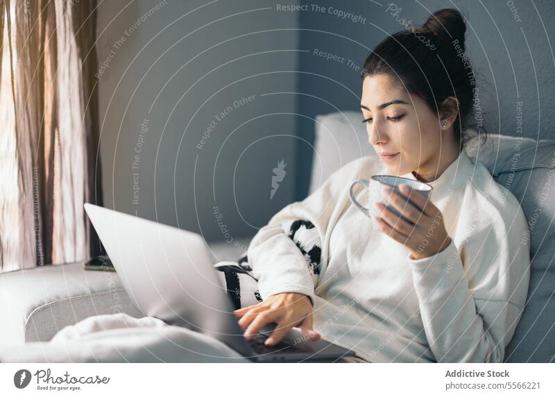 Crop junge Frau sitzt mit Laptop und Kaffeetasse Tasse benutzend Internet Anschluss online Wohnzimmer zu Hause dunkles Haar Sofa Apparatur Gerät Getränk Netbook