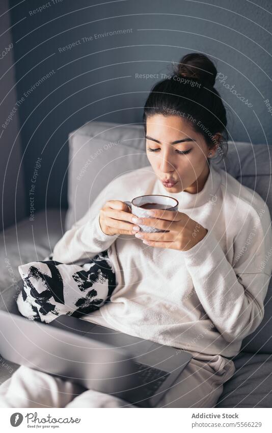 Crop junge Frau sitzt mit Laptop und Kaffeetasse Tasse benutzend Internet Anschluss online Wohnzimmer zu Hause dunkles Haar Sofa Apparatur Gerät Getränk Netbook