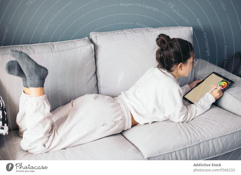 Junge Frau mit Brille liegt auf dem Sofa und surft zu Hause auf einem digitalen Tablet lesen Tablette e-Buch gemütlich Lügen Komfort online Wohnzimmer jung