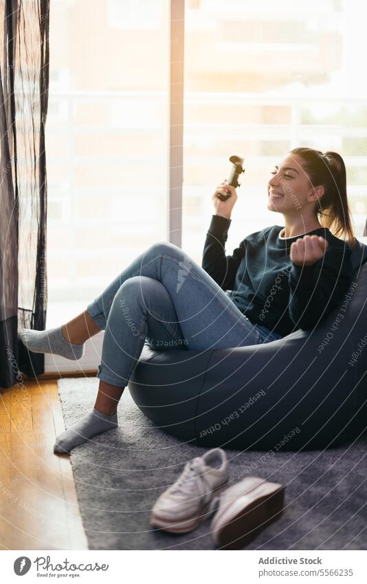 Fröhliche Frau mit Joystick das Spiel zu Hause besinnlich Konsole benutzend Videospiel mit den Achseln zucken gestikulieren Bohnenbeutel heimwärts Gerät jung