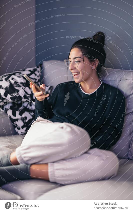 Fröhliche Frau ruht sich auf dem Sofa aus und liest Nachrichten auf ihrem Smartphone Textnachricht lesen Lächeln Glück online Wohnzimmer zu Hause jung Brille
