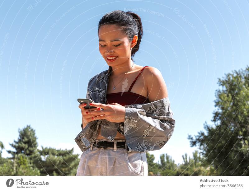 Asiatische Frau im Freien mit Smartphone-Nutzung an einem sonnigen Tag. stylisch Outfit Mobile Telefon Technik & Technologie jung Park Sommer modern lässig