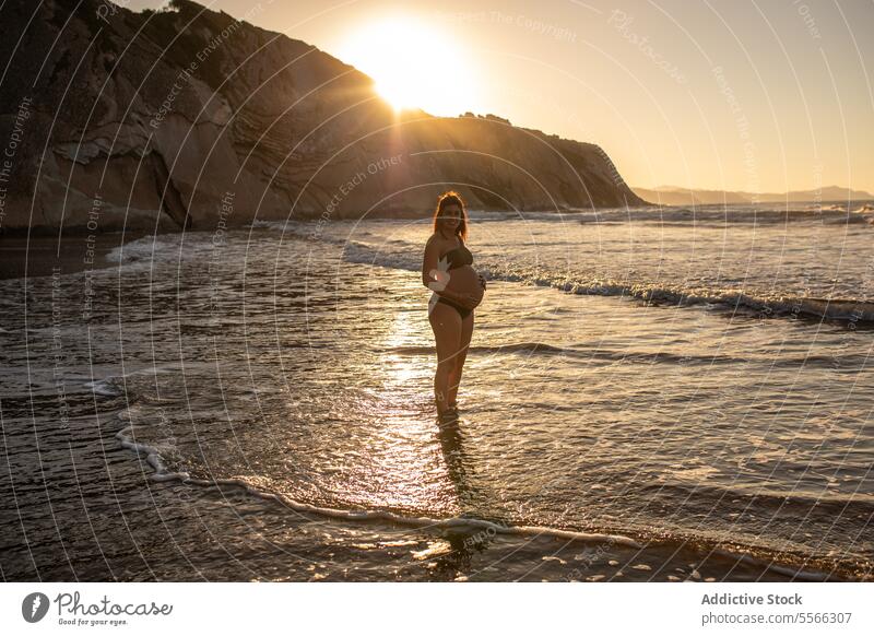 Lächelnde schwangere Frau im Meerwasser stehend Bauch erwarten MEER Bikini Sommer Sonnenuntergang berühren Glück heiter pränatal vorwegnehmen Schwangerschaft