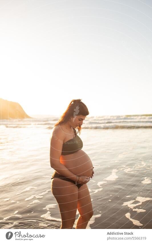 Schwangere Frau im Meerwasser stehend schwanger Bauch erwarten MEER Bikini Sommer Sonnenuntergang berühren Glück heiter Lächeln pränatal vorwegnehmen