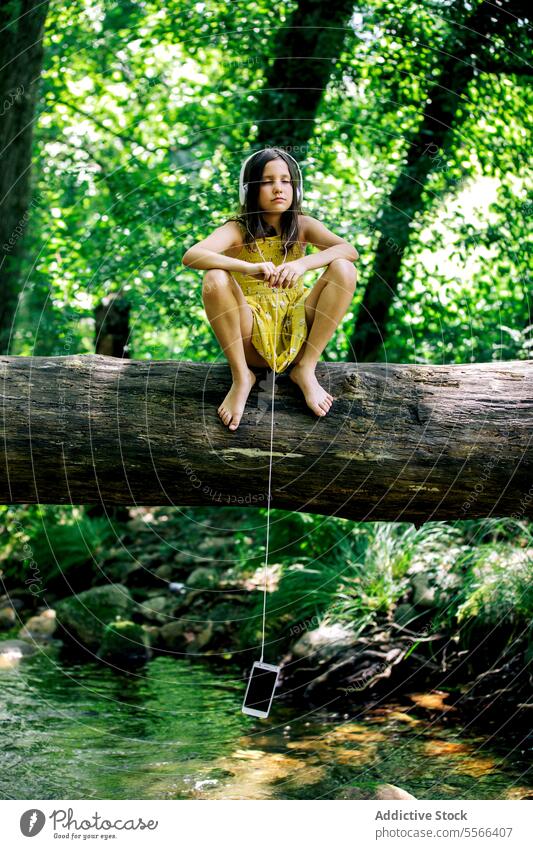 Mädchen im Teenageralter sitzt auf einem Baumstamm und hört Musik über Kopfhörer mit hängendem Mobiltelefon Wald Kofferraum Pause Erholung Natur Abenteuer ruhig